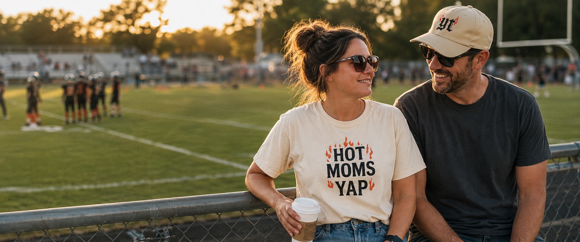 A mom in a cream YapThreads Hot Moms Yap tee and a dad in a YapThreads yt cap smiling together at a football sideline at sunset