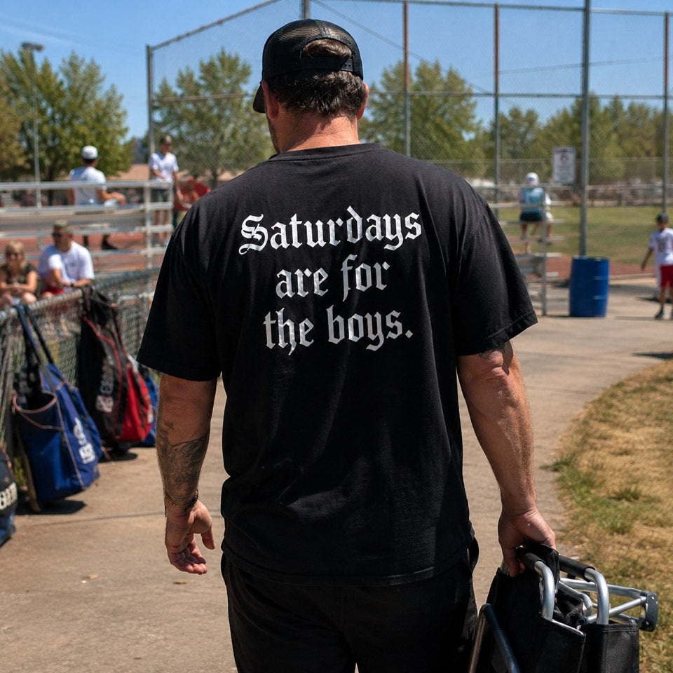 Dad walking up to the ballfield in the black Saturdays Are For The Boys tee, carrying a folding chair