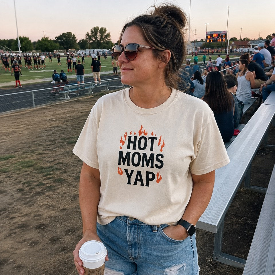 Mom in sunglasses on the football sideline at dusk wearing the cream Hot Moms Yap tee and holding a coffee
